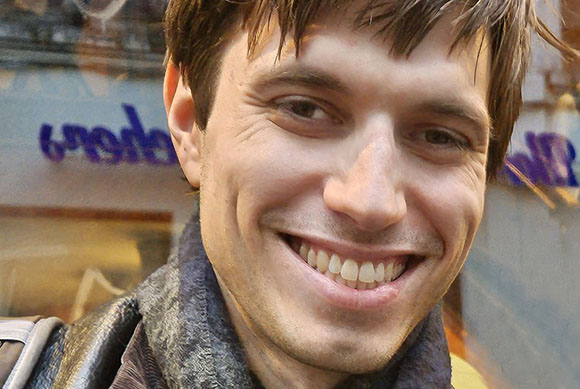 A close-up portrait of a smiling man with brown hair and a patterned scarf.