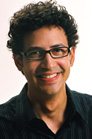 A smiling man with dark curly hair and glasses wearing a black button-down shirt.