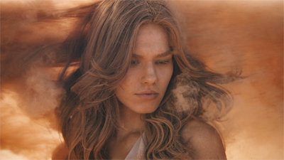A woman with long wavy hair looking forward through swirling orange dust and sand.