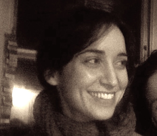 A smiling woman with dark hair wearing a thick scarf in a dimly lit room.