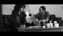 Two men smiling and sitting on a leather couch next to an acoustic guitar in a black and white photo.