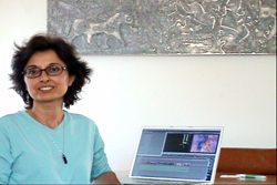 A woman with short dark hair and glasses wearing a light blue shirt sits at a desk with an open laptop.