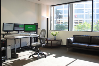 An office with a computer desk, an ergonomic chair, a small coffee table with flowers, and a black leather sofa.