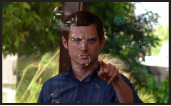 A man in a blue shirt points toward the camera as iridescent bubbles float around him.