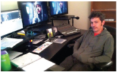 A man sits in an office chair at a desk with three computer monitors and editing equipment.