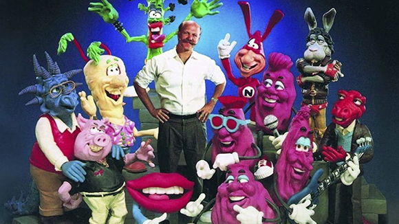 A man in a white shirt stands surrounded by many colorful, expressive puppet characters.