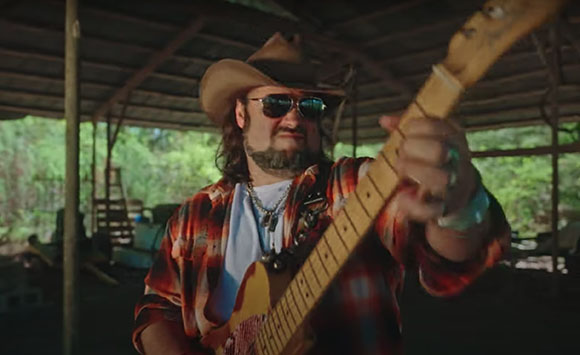 A man wearing a cowboy hat and sunglasses plays an electric guitar under a wooden shelter.