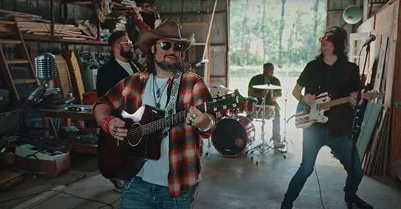 A band playing acoustic guitar, electric guitar, and drums inside a cluttered garage.