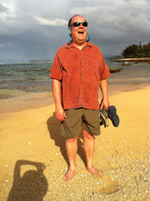 A man in an orange shirt and sunglasses laughs while standing on a sandy beach under a cloudy sky.