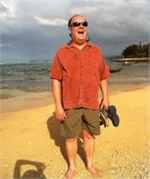 A man in an orange shirt and sunglasses laughs while standing on a sandy beach.