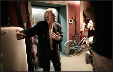 A man in a black blazer gestures with both hands in a room containing an elevator and a blue bicycle.