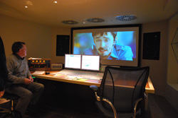 A person sits at a desk in a dark room, watching a man on a large computer monitor.