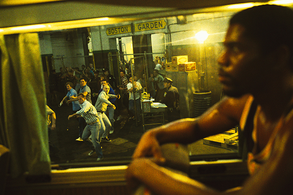 People gathered in a room with a sign reading Boston Garden, seen through a window from a dark interior.