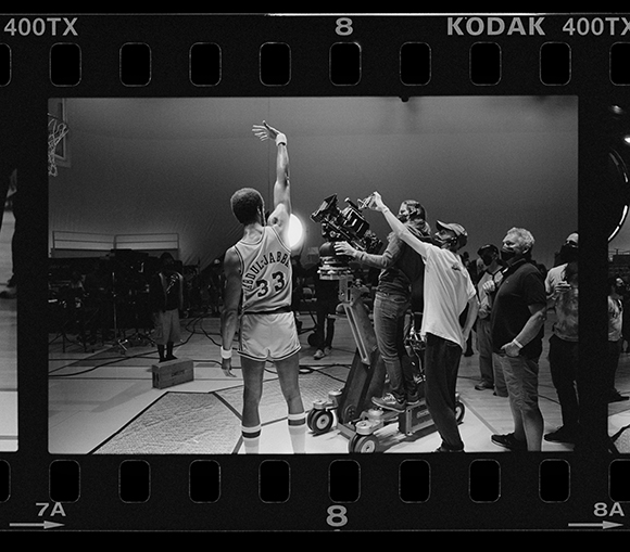 A basketball player reaches up while a film crew operates a large camera on a dolly in a gym.