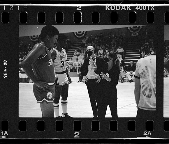 Black and white film frame of basketball players and staff standing on a court during a game.