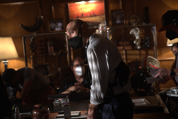 A man in a white shirt and dark mask looks downward in a dimly lit, cluttered room.