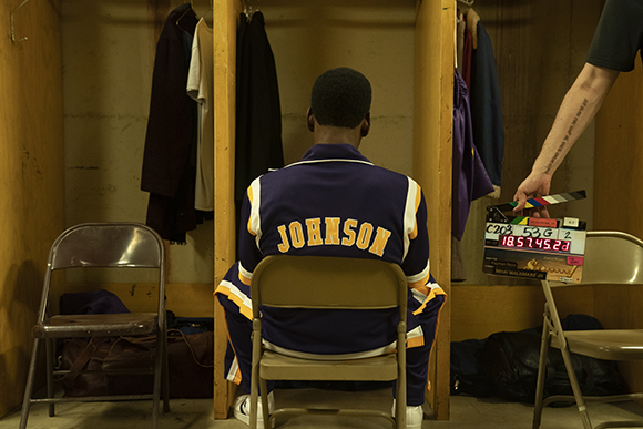 A person in a purple and yellow Johnson varsity jacket sits on a chair facing away in a locker room.