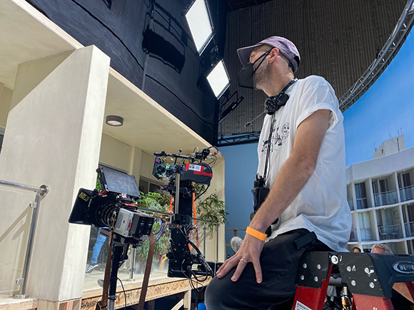 A man in a white t-shirt and face mask sits near a large professional film camera on a production set.
