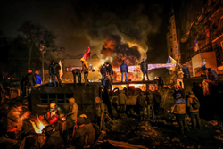 Protesters stand near fires and a large vehicle amidst smoke at night.
