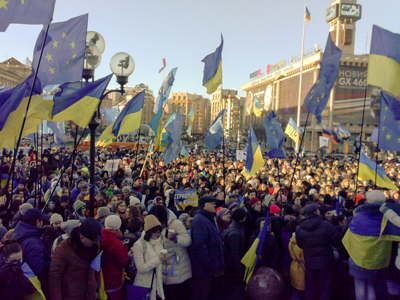 A large crowd of people gathers in a city square, waving many blue and yellow flags under a bright sky.