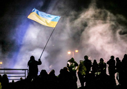 Silhouette of a person holding a blue and yellow Ukrainian flag aloft amidst smoke and a crowd at night.