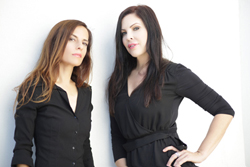 Two women with long hair wearing black tops pose against a plain white wall.