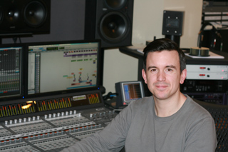A man in a grey shirt sits behind a large mixing console in a professional recording studio.