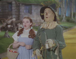 A young woman with red hair looks surprised next to a scarecrow in a green outfit.