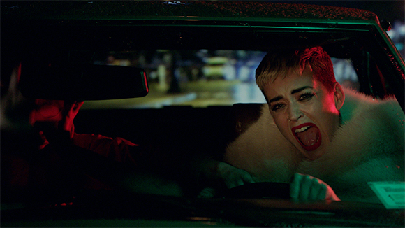 A woman with short red hair and a white fur coat screams inside a car at night.