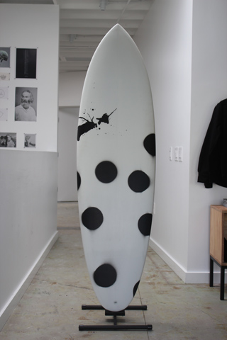A white surfboard with large black polka dots and a black silhouette design stands on a rack in a white hallway.
