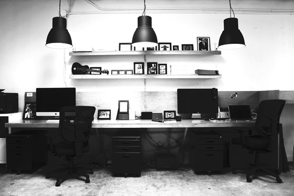 Two workstations with monitors, chairs, and filing cabinets under two long shelves and three hanging lamps.
