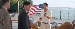 Three men in business and casual attire converse on a boat deck with an American flag in the background.