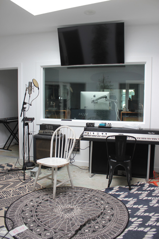 A recording studio featuring a piano, microphone, chair, and a large window looking into a control room.