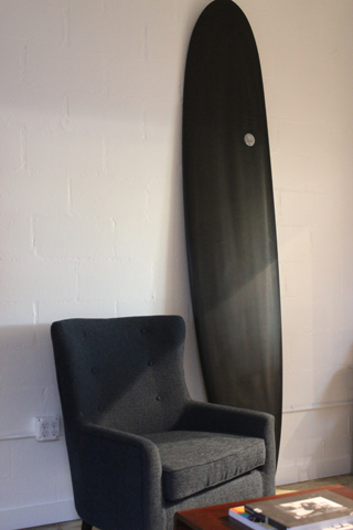 A long black surfboard leans against a white brick wall behind a grey upholstered armchair.