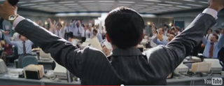 A man in a dark pinstripe suit raises both arms in a large, busy office filled with people at desks.