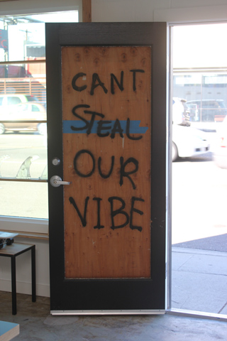 A black door with a wooden center panel featuring the handwritten text 'CANT STEAL OUR VIBE' in black ink.