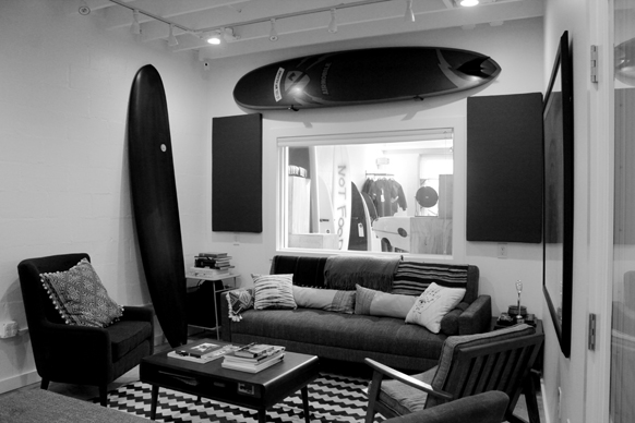 Black and white photo of a living room with a sofa, armchairs, a coffee table, and a surfboard on the wall.