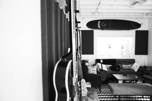 Black and white photo of guitars in the foreground and a lounge area with a surfboard on the wall in the background.