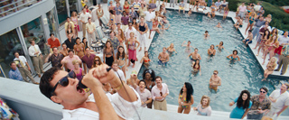 A man sings into a microphone in the foreground of a large, crowded outdoor pool party.