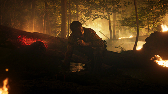 A man crouches in a dark, smoky forest near glowing embers and small fires.