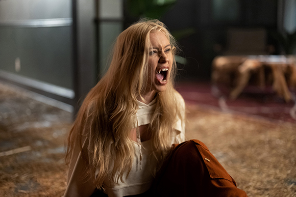 A blonde woman with long hair and vampire fangs screams while sitting on a floor.