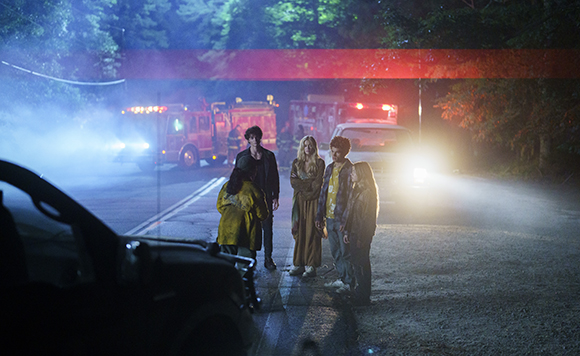 Five young people stand on a dark road at night near a fire truck and a car with bright headlights.
