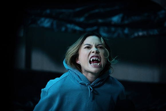 A woman with long fangs and a snarling expression wears a blue hoodie in a dark setting.