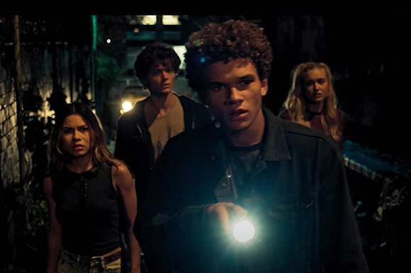 Four young adults walk through a dark, wooded area at night, one holding a bright flashlight.