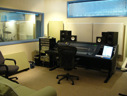 A music studio control room with a large mixing console, studio monitors, and an ergonomic desk chair.