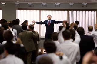 A man in a dark suit stands with arms wide in a room filled with people facing him.