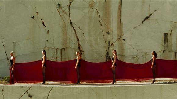 Five women in black outfits hold a long, flowing red fabric in front of a large, textured stone cliff.