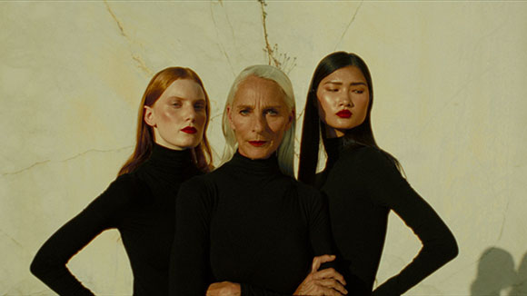 Three women with different hair colors and features wearing black turtlenecks against a light marble background.