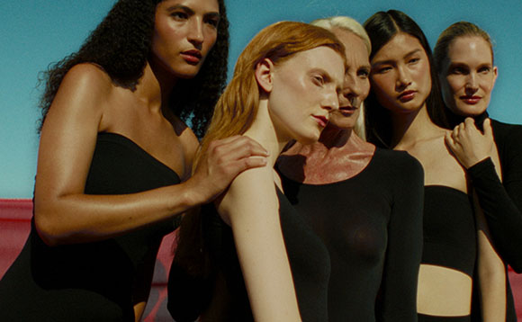 Five diverse models in black outfits pose closely together against a bright blue sky.