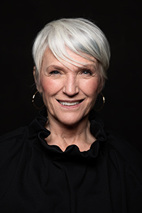 A smiling woman with short white hair and gold hoop earrings wearing a black ruffled shirt against a black background.